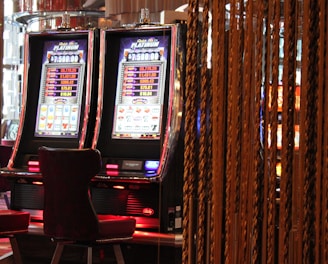 A pair of casino slot machines, each with detailed screens displaying various game outcomes and jackpot amounts. The chairs in front of the slot machines are dark and cushioned for comfort. The background features a hanging curtain made of golden ropes and other casino decorations are visible.