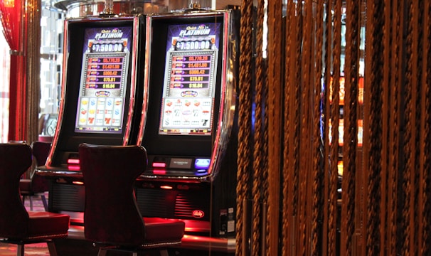 A pair of casino slot machines, each with detailed screens displaying various game outcomes and jackpot amounts. The chairs in front of the slot machines are dark and cushioned for comfort. The background features a hanging curtain made of golden ropes and other casino decorations are visible.