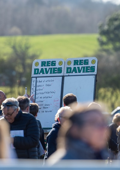 A group of people gathered outdoors with two large betting boards in the background labeled 'REG DAVIES'. The setting appears to be a countryside location with trees and fields visible in the distance. The crowd consists of individuals dressed in jackets and hats, engaging in conversation.