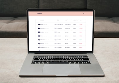 A laptop on a table displaying a cryptocurrency trading platform. The screen lists various cryptocurrencies along with their buy and sell values, spread percentages, and 24-hour change percentages. The keyboard and touchpad of the laptop are visible, set against a neutral background with a sofa in the back.