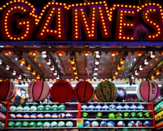 A brightly lit carnival game stall with a large 'GAMES' sign at the top surrounded by red and yellow lights. Various sports balls, including basketballs and soccer balls, are hanging from the ceiling and neatly arranged on shelves below.