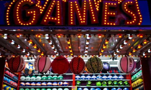A brightly lit carnival game stall with a large 'GAMES' sign at the top surrounded by red and yellow lights. Various sports balls, including basketballs and soccer balls, are hanging from the ceiling and neatly arranged on shelves below.